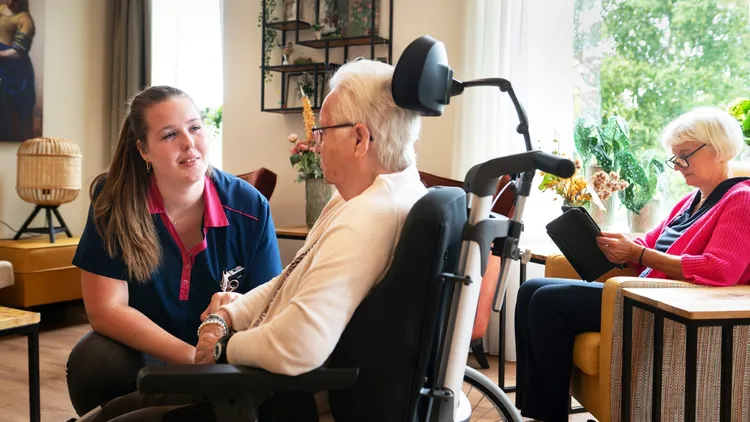Huiskamer met zorgmedewerker en bewoner in rolstoel foto Niels Zomer